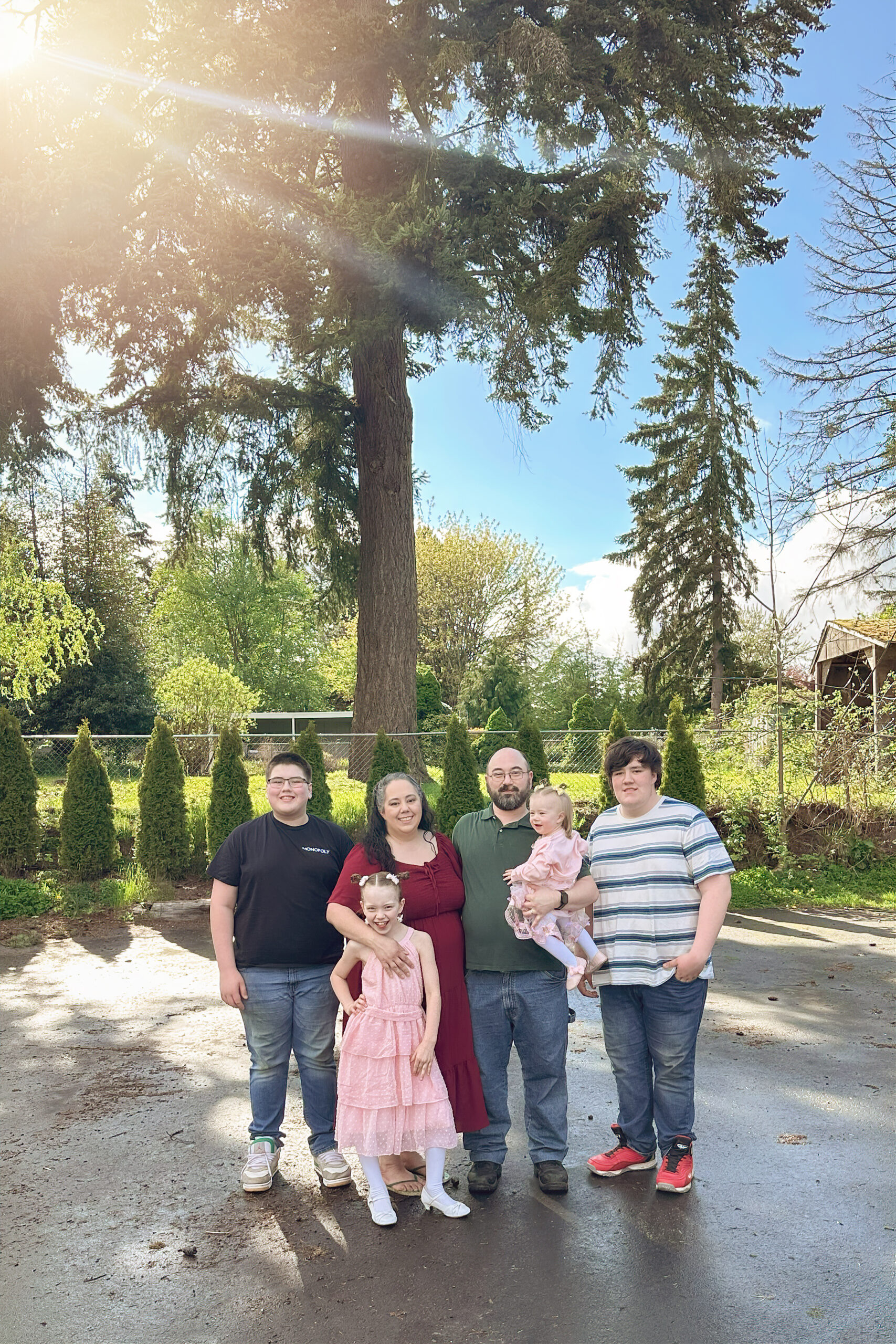 Family of six standing outdoors in spring sunlight, smiling together with tall evergreen trees in the background in Puyallup, WA.